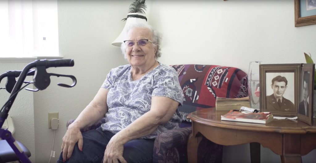 Older woman seated on couch surrounded by family photos smiles; walker is parked nearby