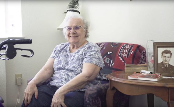 Older woman seated on couch surrounded by family photos smiles; walker is parked nearby