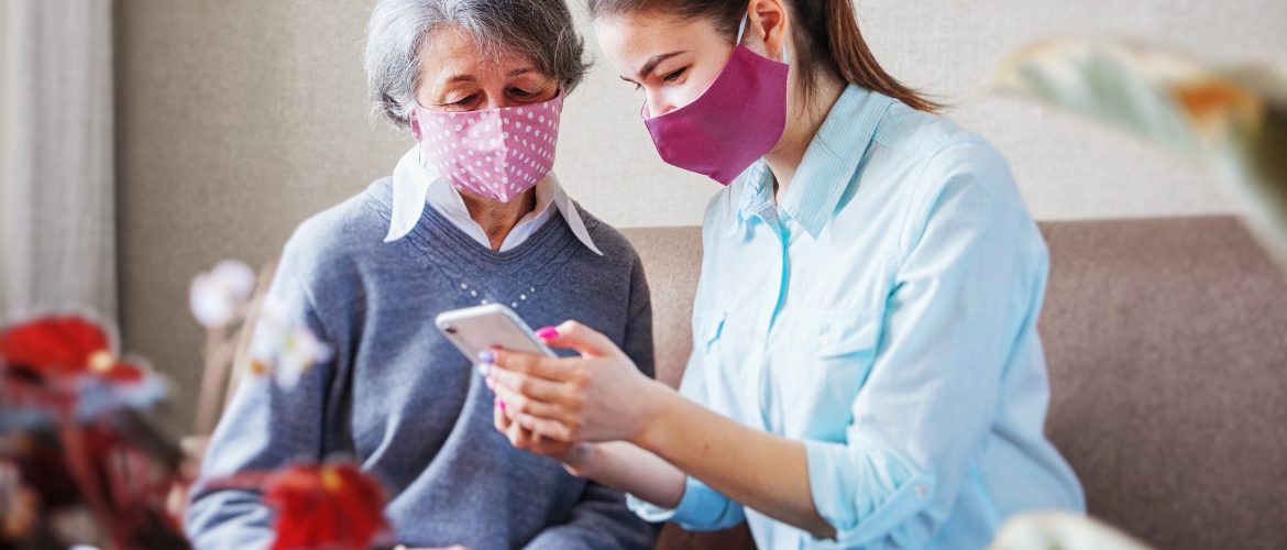 A younger woman wearning a mask shows something on her phone to an older woman wearing a mask