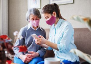 A younger woman wearning a mask shows something on her phone to an older woman wearing a mask