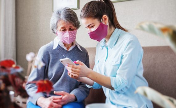 A younger woman wearning a mask shows something on her phone to an older woman wearing a mask
