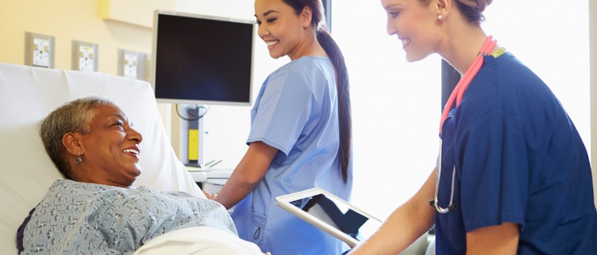 Smiling Nurse With Digital Tablet Talks To Female Patient In Hospital Bed