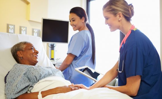 Smiling Nurse With Digital Tablet Talks To Female Patient In Hospital Bed