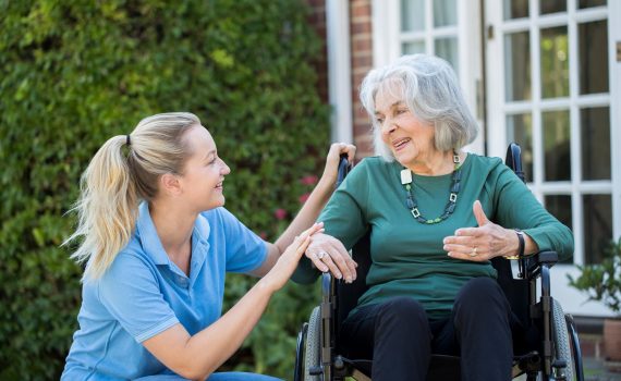 Carer Pushing Senior Woman In Wheelchair Outside Home