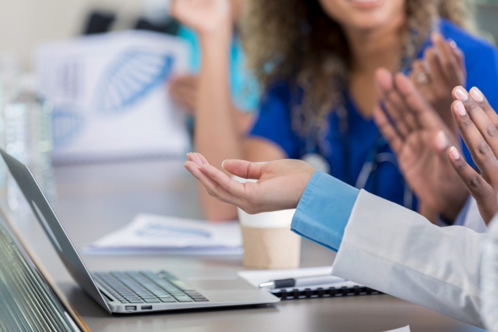 A laptop is on the table along with conference materials. Point-of-care workers are talking to each other at a conference table.