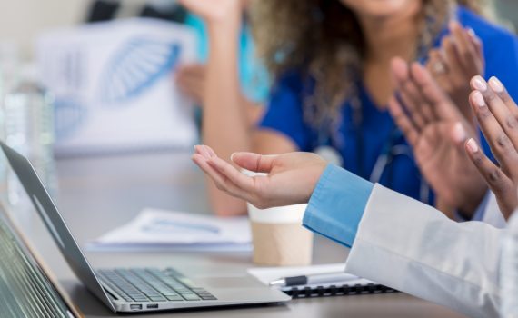A laptop is on the table along with conference materials. Point-of-care workers are talking to each other at a conference table.