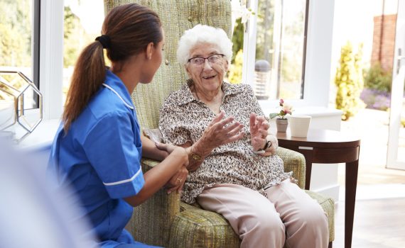 Senior Woman Sitting In Chair And Talking With Nurse In Retirement Home