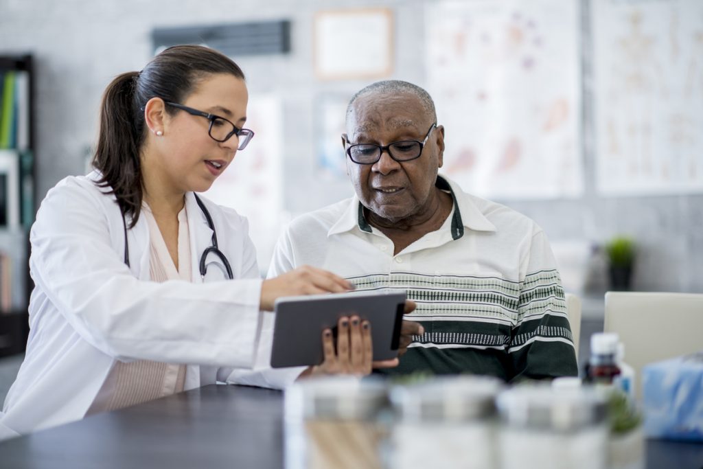 A senior male is sitting with a female doctor who is showing him something on a tablet