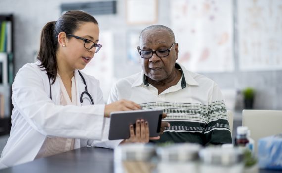 A senior male is sitting with a female doctor who is showing him something on a tablet