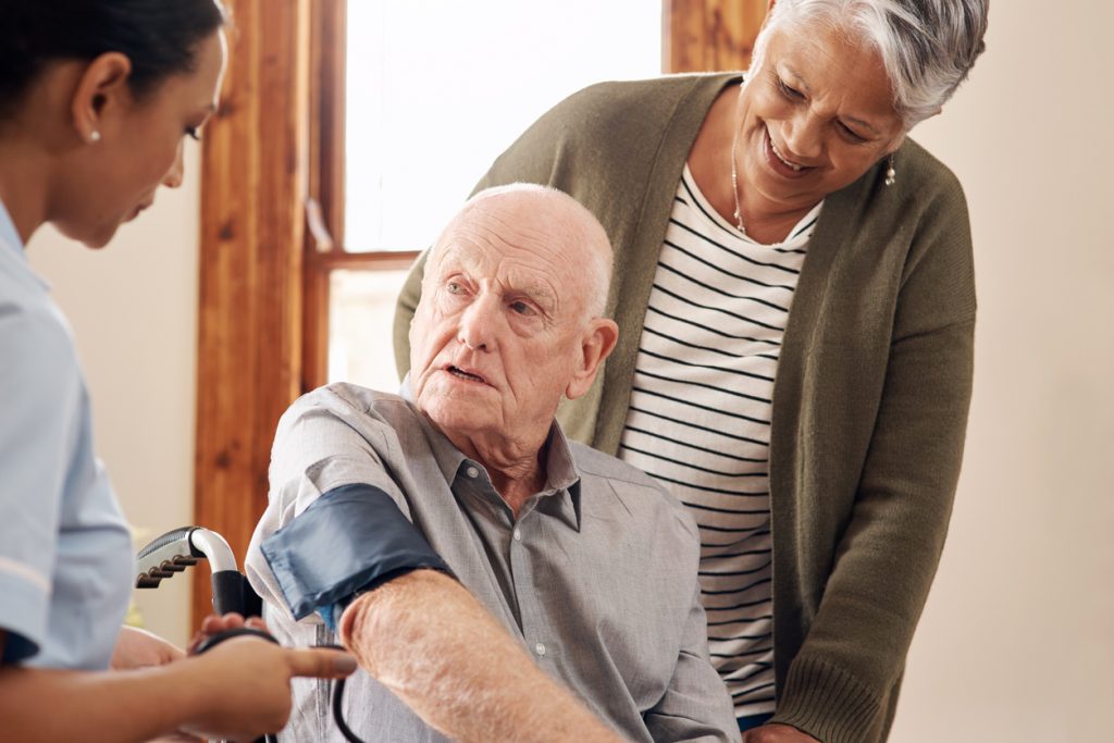 Senior man uses blood pressure cuff while point-of-care worker and senior woman look on