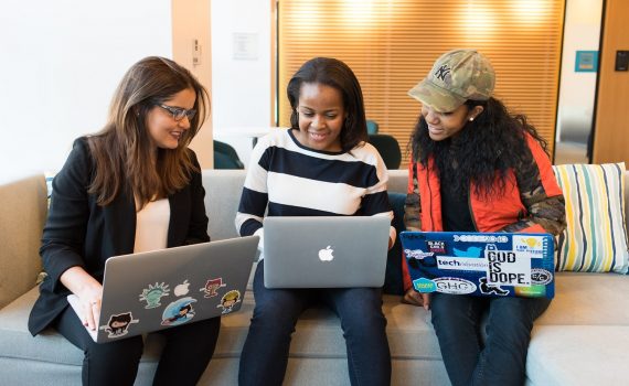 Three women sitting on a couch looking at laptops