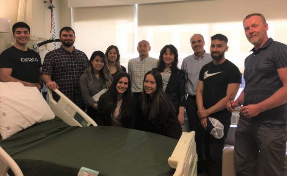 11 people standing around a hospital bed smiling.